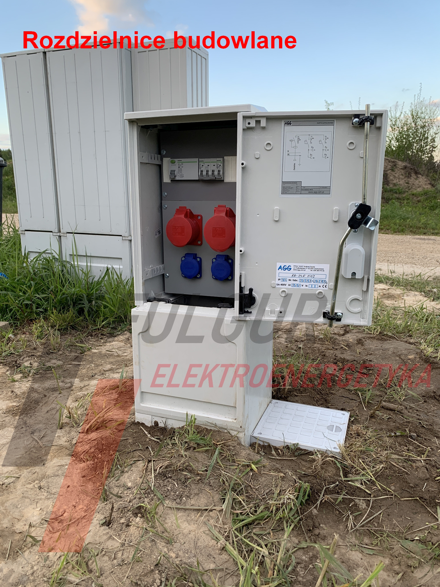 Otwarta, biała rozdzielnica budowlana AGG z czerwonymi i niebieskimi gniazdami, umieszczona na ziemi z trawą. Widoczna karta katalogowa i inne rozdzielnice w tle.