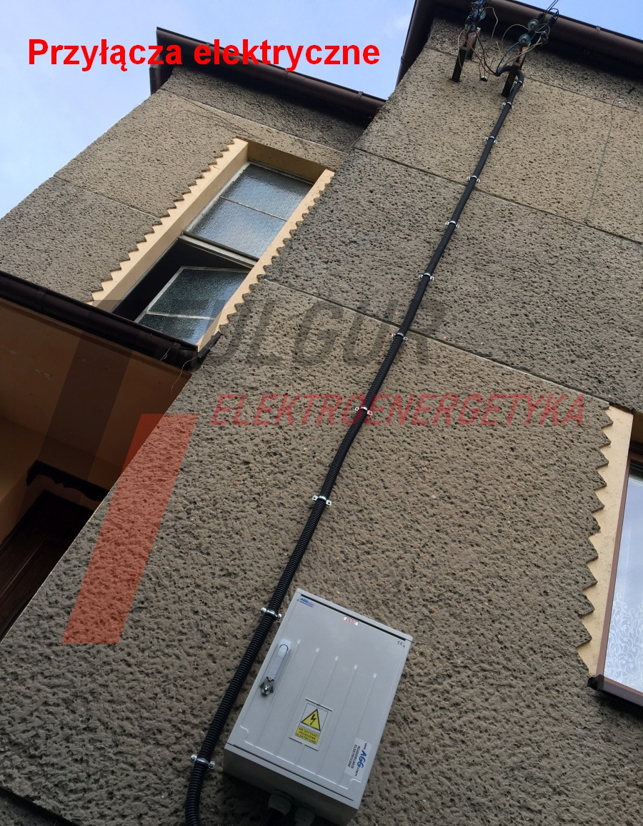 Zewnętrzna instalacja elektryczna na elewacji budynku z szarą skrzynką i czarnym kablem w peszlu, mocowanym klamrami. Widok z dołu, lekki kąt.