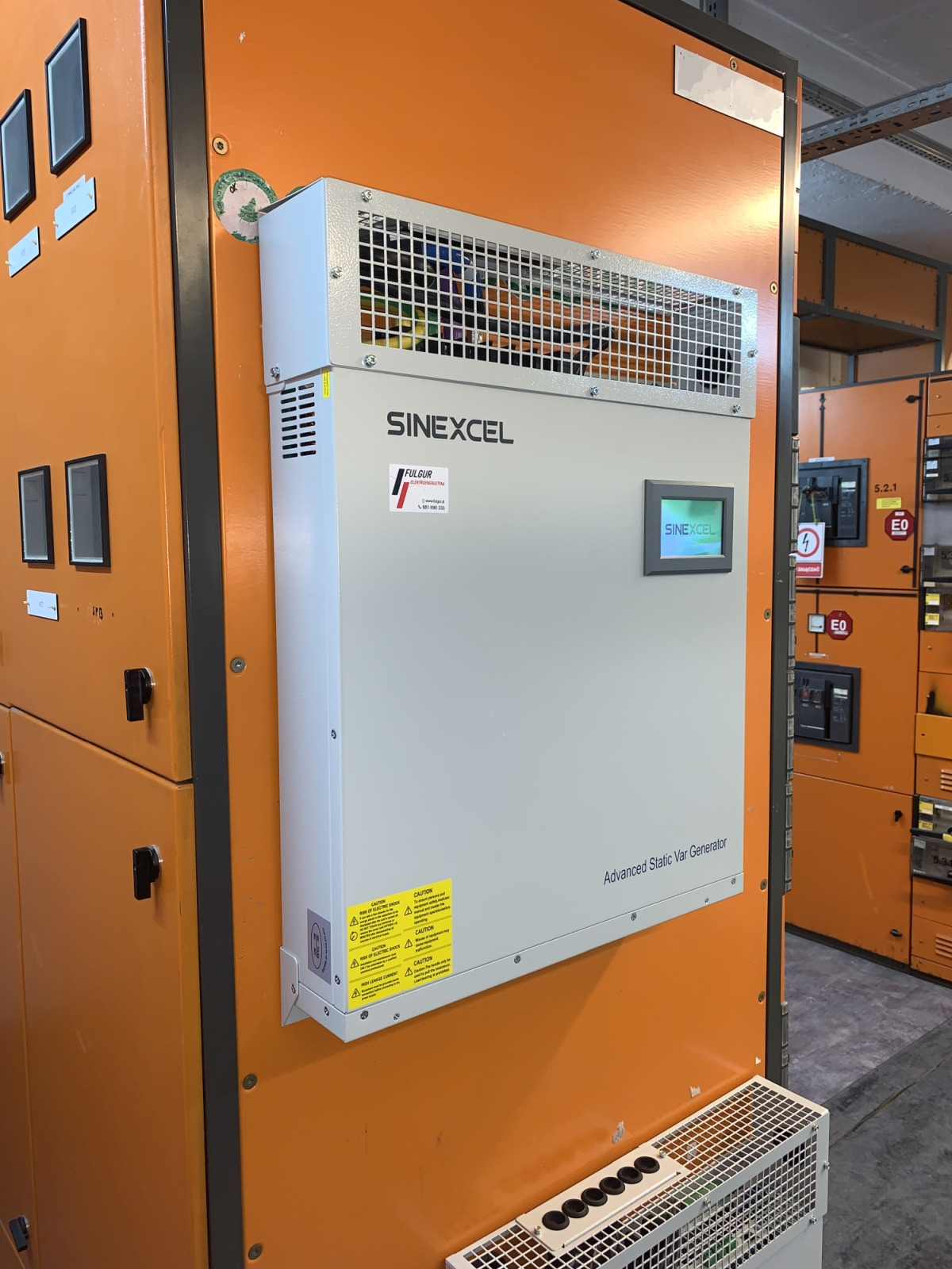 Szaro-biały kompensator mocy biernej Sinexcel zamontowany na pomarańczowej szafie elektrycznej. Widoczne ostrzeżenia o napięciu i ekran LCD z logo Sinexcel.