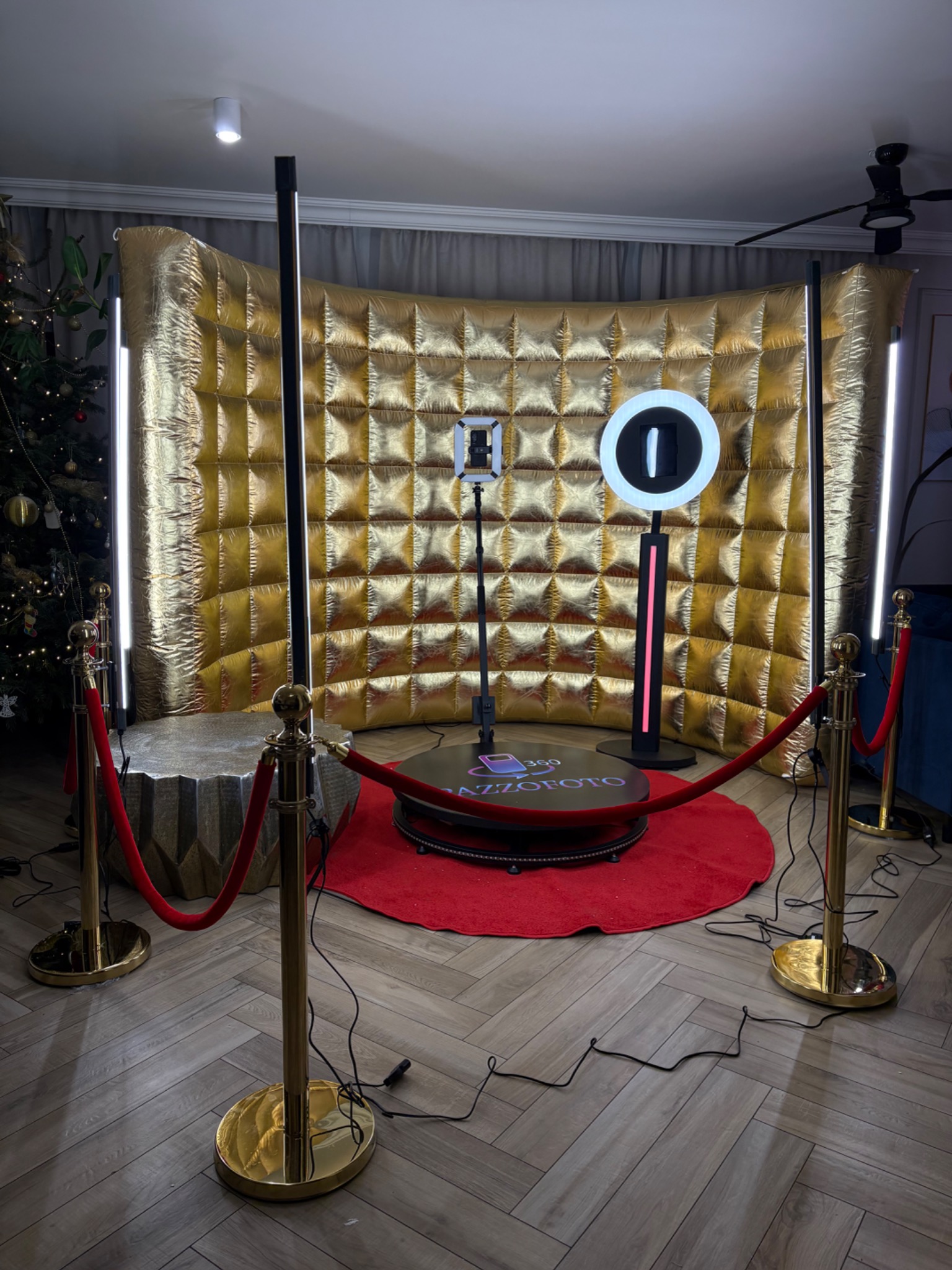 Złota ścianka, czerwony dywan i złote słupki z czerwonym sznurem tworzą elegancką oprawę fotobudki 360. Świąteczny klimat podkreśla choinka w tle.