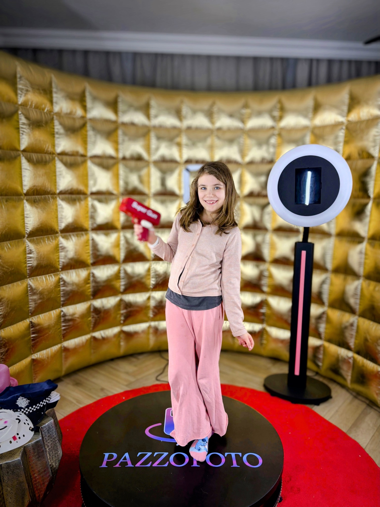 Uśmiechnięta dziewczynka z rekwizytem w dłoni pozuje w fotobudce z błyszczącym, złotym tłem. Na pierwszym planie widoczna platforma z logo 'PAZZOFOTO' i rekwizyty.