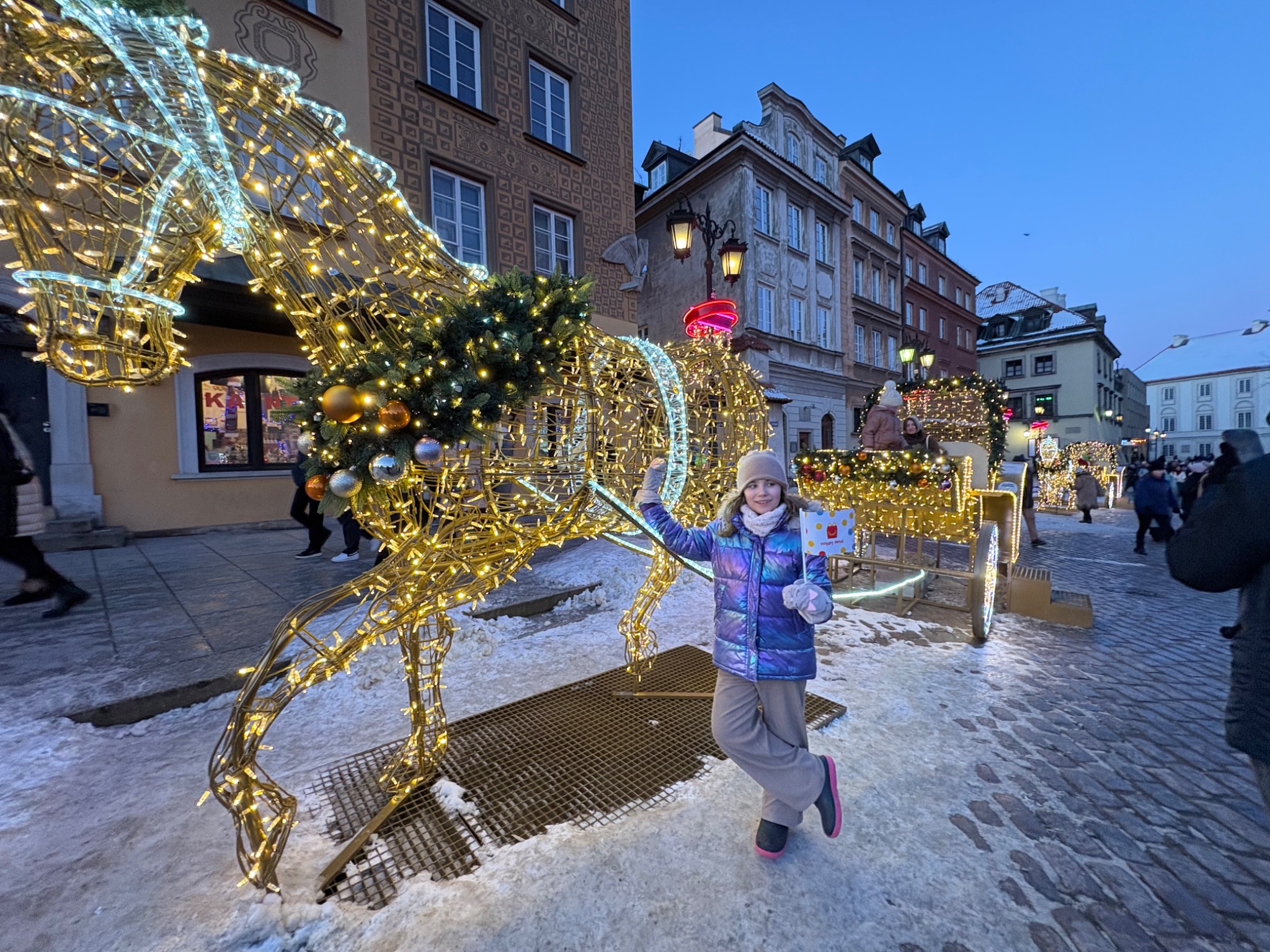 Dziewczynka pozuje przy świątecznej iluminacji konia i karocy na tle architektury Starego Miasta w Warszawie. Zimowa sceneria, wieczorne oświetlenie.