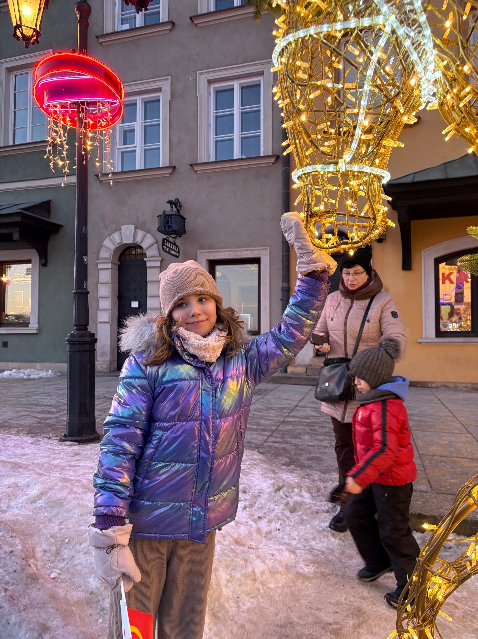 Dziewczynka w błyszczącej kurtce pozuje na tle świątecznych dekoracji w zimowej scenerii miejskiej. W tle budynki i inne osoby.