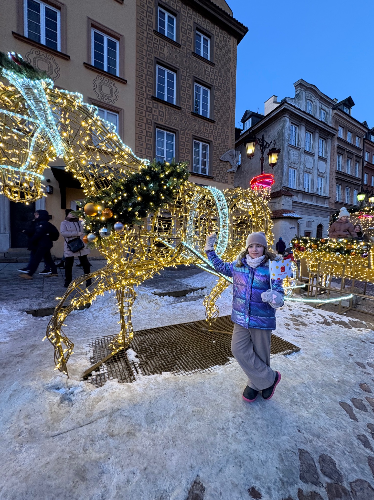 Dziewczynka na tle świetlnej dekoracji konia i karocy na tle staromiejskich kamienic w zimowej scenerii. Dekoracja świąteczna z lampkami.
