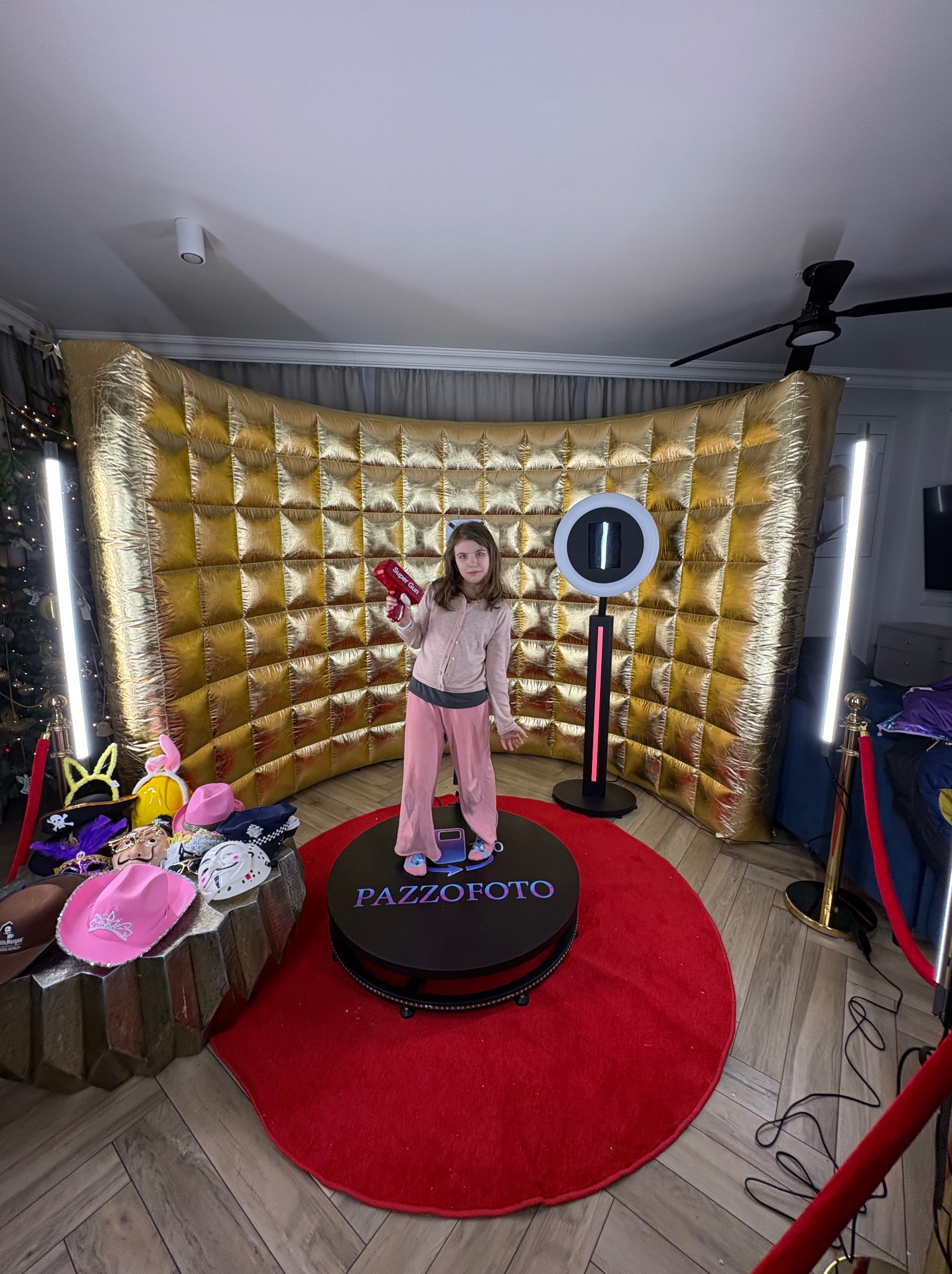 Dziewczynka z rekwizytem na obrotowej platformie fotobudki 360, na czerwonym dywanie, z złotym tłem i oświetleniem. W tle rekwizyty do zdjęć.