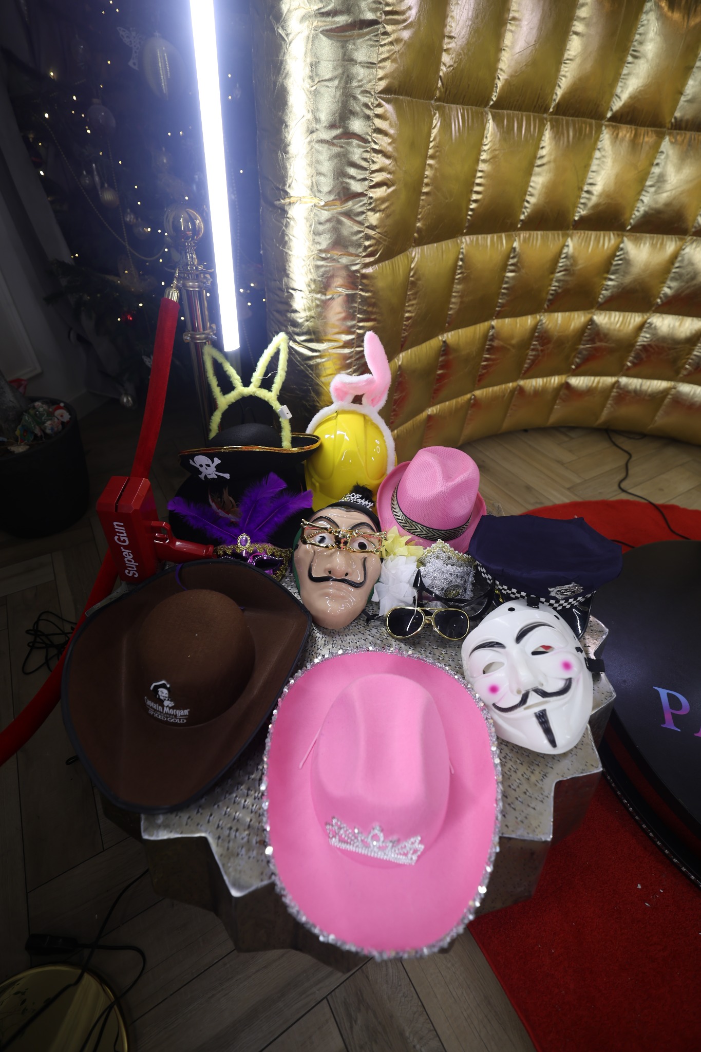 Różnorodne rekwizyty fotobudki: kapelusze kowbojskie, maski anonima i Salvadora Dali, uszy królika, piracki kapelusz, okulary, na tle złotej ściany.