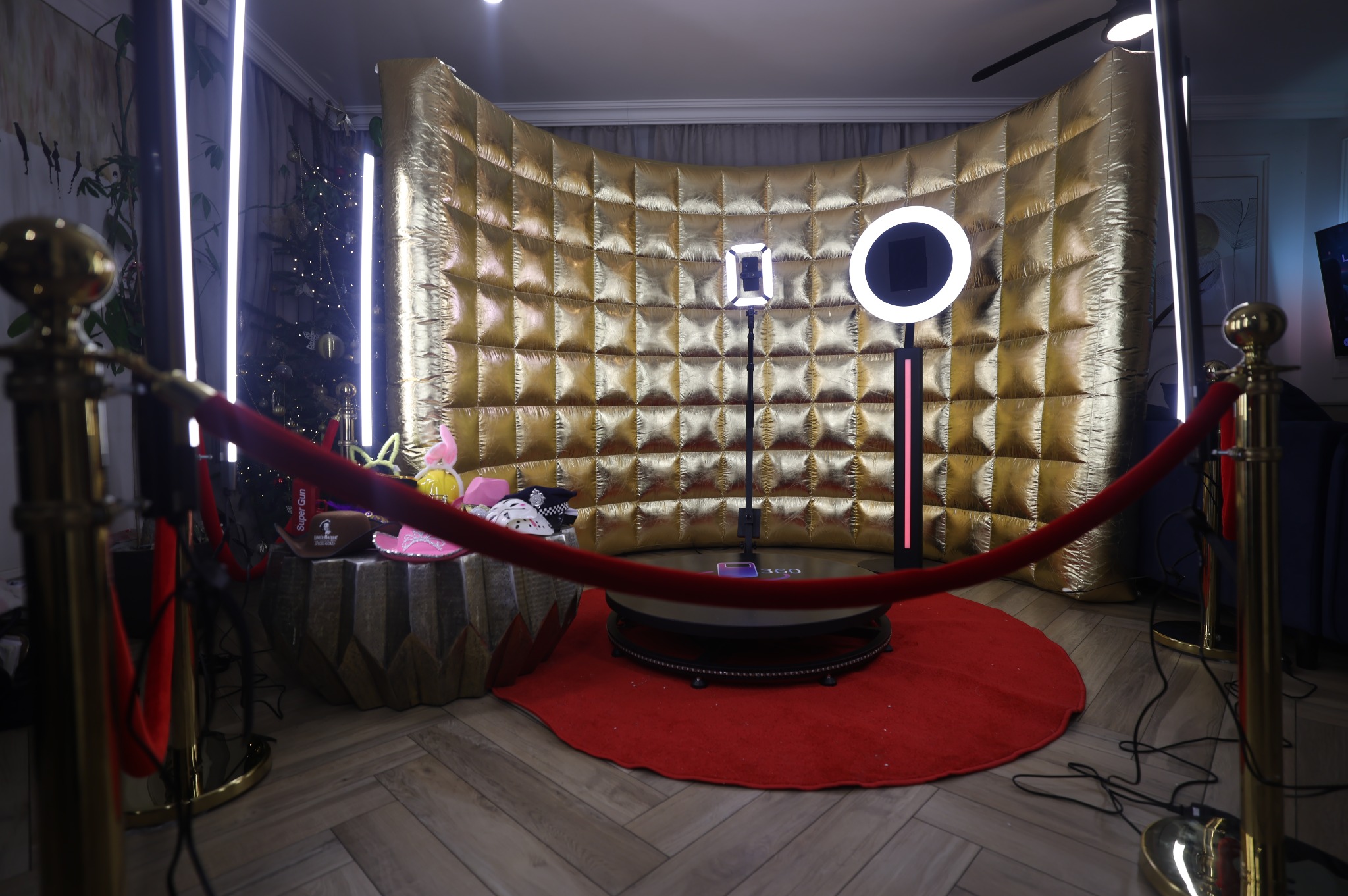 Złota ścianka z podświetleniem LED i rekwizytami na podeście z czerwonym dywanem, odgrodzona ozdobnym słupkiem z czerwonym sznurem. Dekoracja imprezowa.