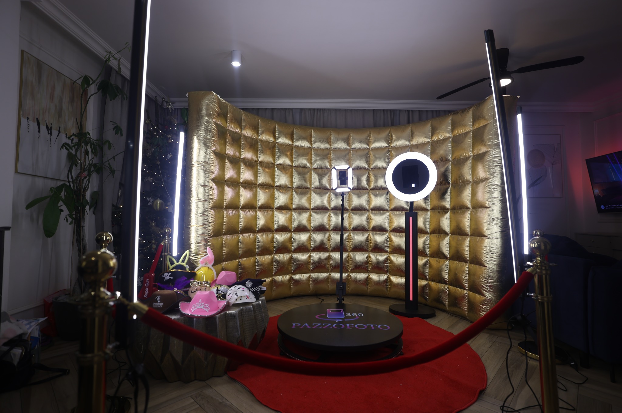 Złota, dmuchana ścianka fotobudki 360 z oświetleniem LED, rekwizytami i czerwonym dywanem. Widoczne elementy dekoracyjne w tle, takie jak roślina i obraz.