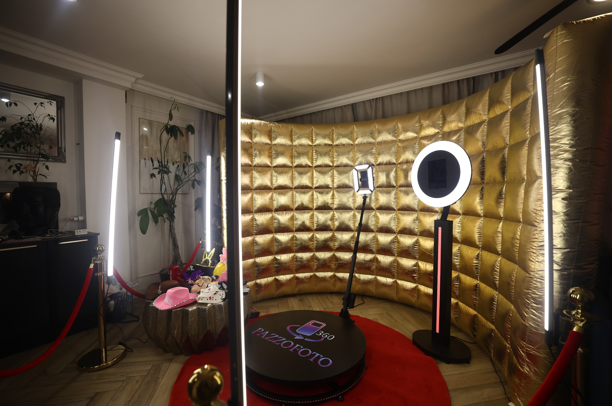 Złota fotobudka 360 z oświetleniem LED i rekwizytami na stoliku, otoczona czerwonym dywanem i słupkami. Widok na wnętrze z dekoracjami i roślinami.