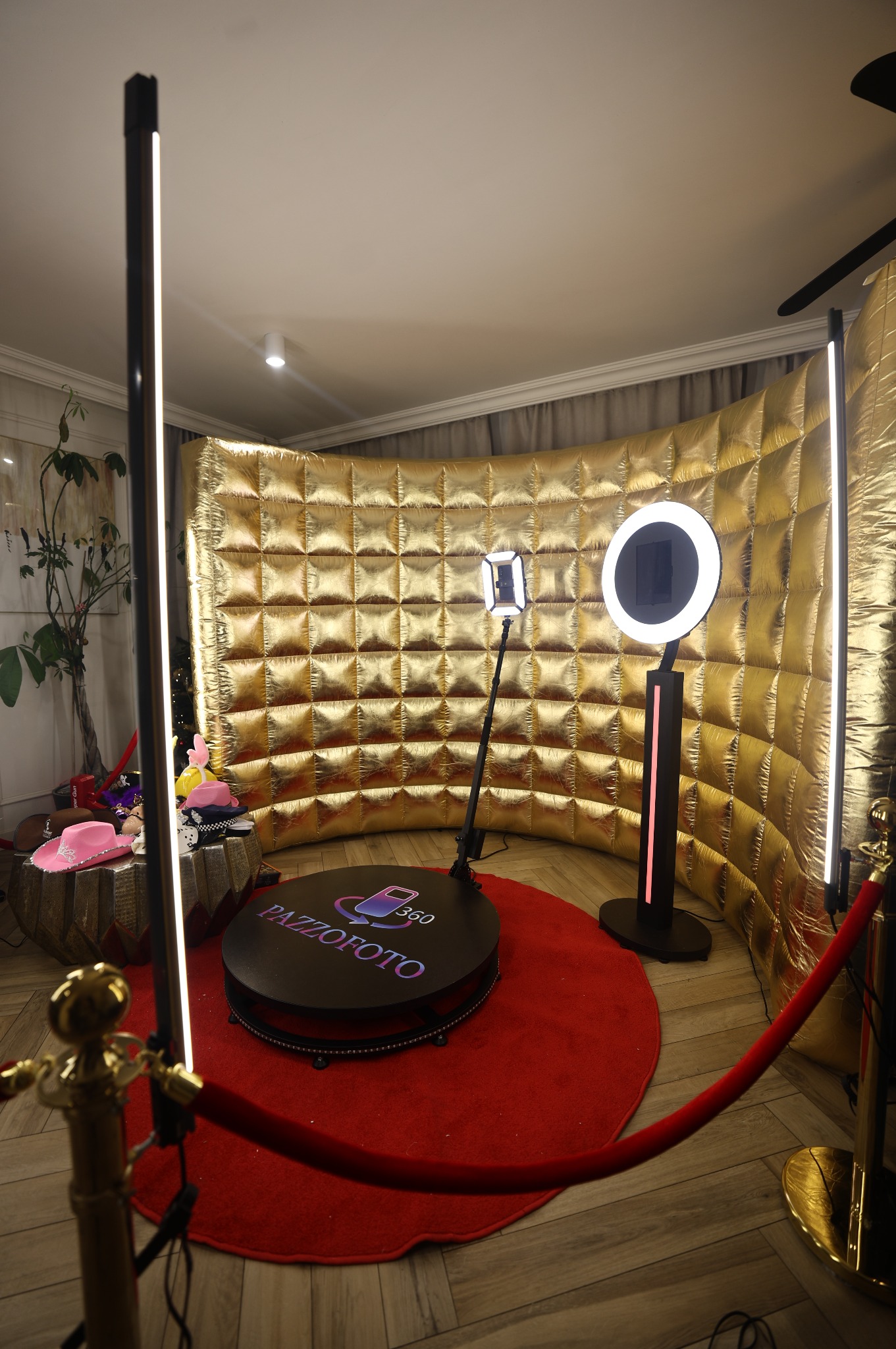 Złota ścianka, czerwony dywan i obrotowa platforma z logo 'PAZZOFOTO 360' tworzą elegancką przestrzeń fotobudki na imprezy. Dwa rodzaje lamp doświetlają scenę.