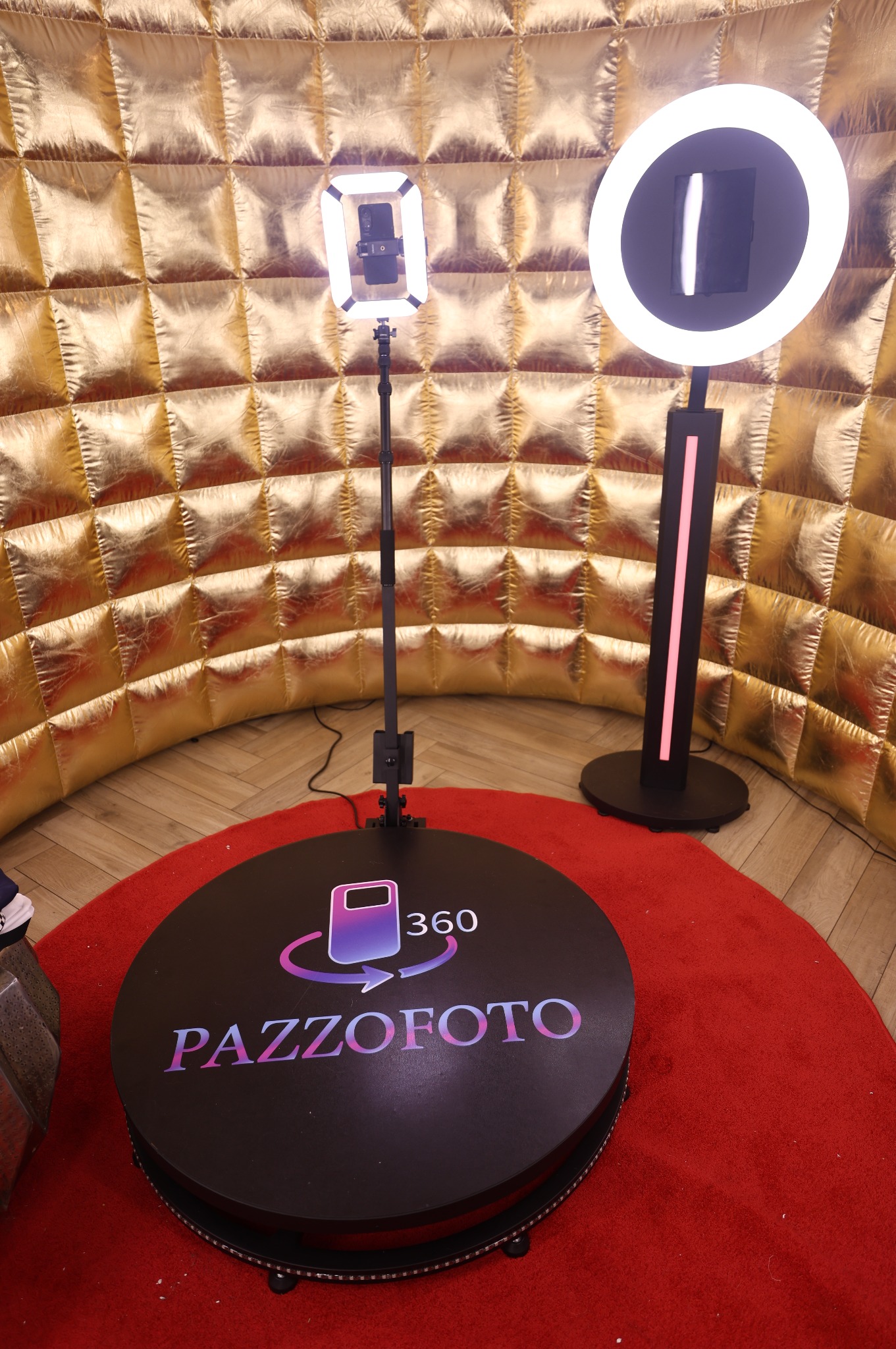 Oświetlona fotobudka 360 z logo PAZZOFOTO na czerwonym dywanie i złotym tle. Dwa różne rodzaje lamp oświetleniowych. Sprzęt gotowy do użycia.