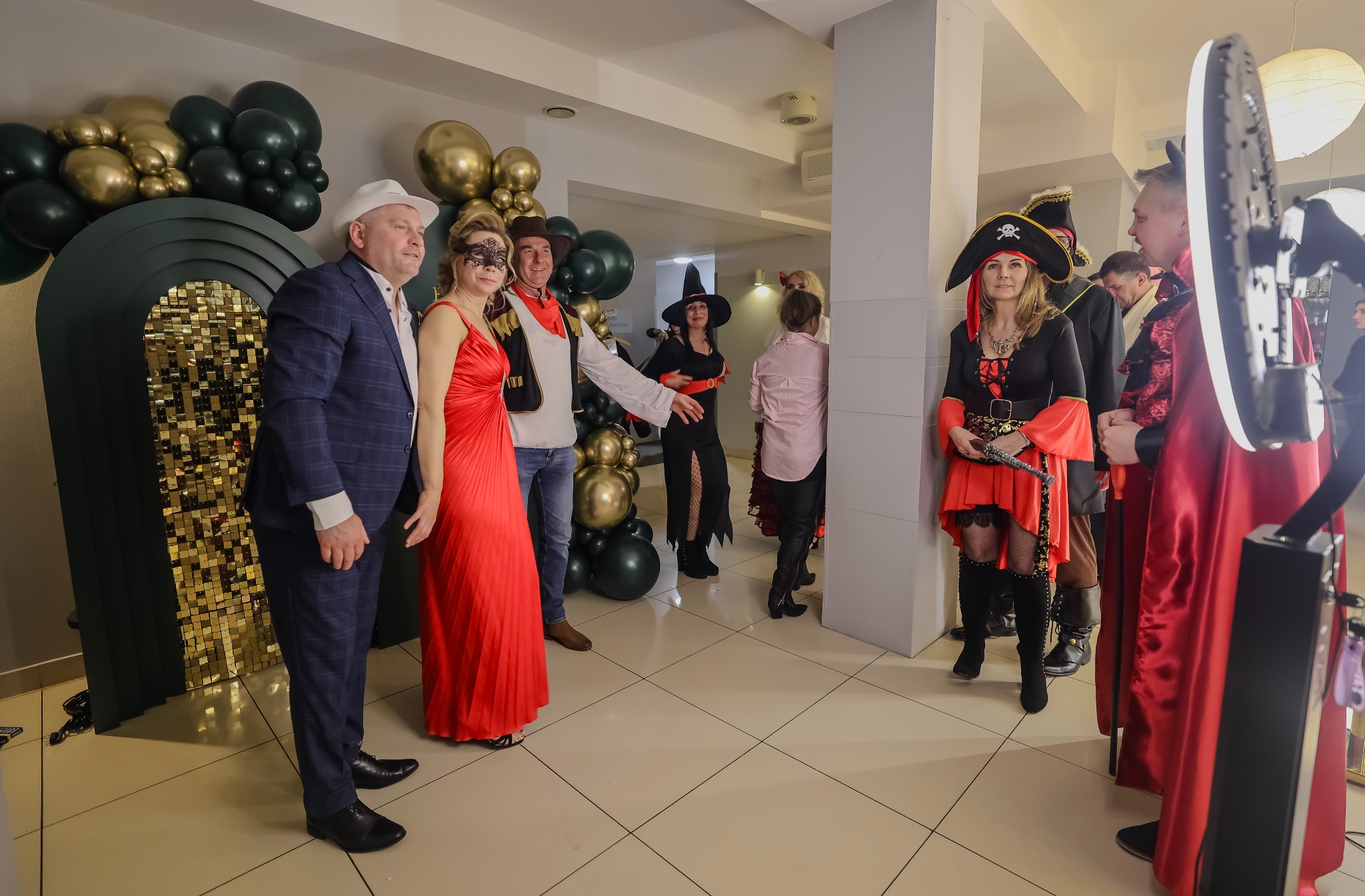 Grupa osób w przebraniach pozuje do zdjęcia przy fotobudce na tle dekoracji z balonów. Widoczny mężczyzna w garniturze i kobieta w czerwonej sukni.