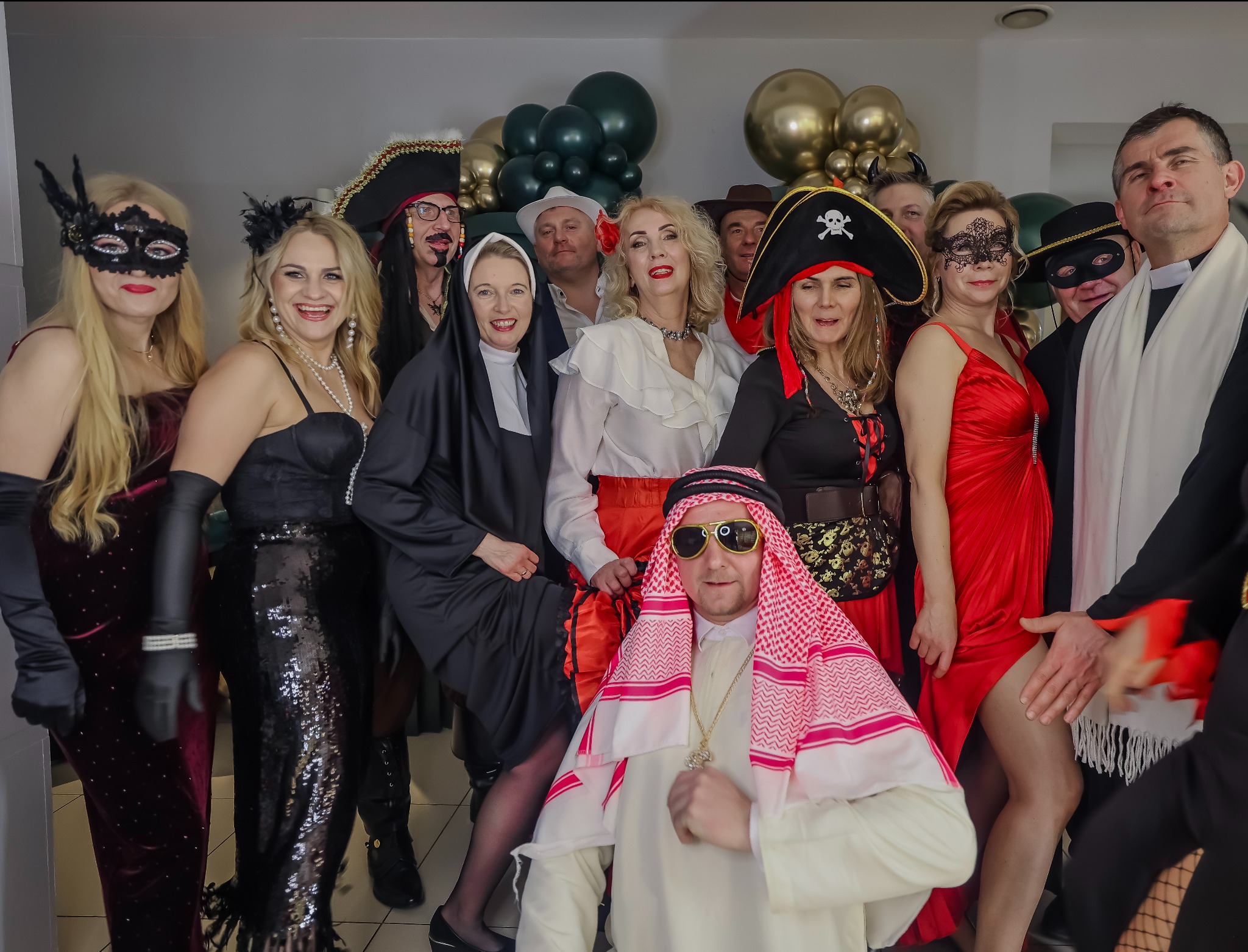Grupa osób w różnorodnych kostiumach karnawałowych, w tym pirat, szejk, zakonnica i postacie w maskach, pozuje na tle dekoracji z balonów.