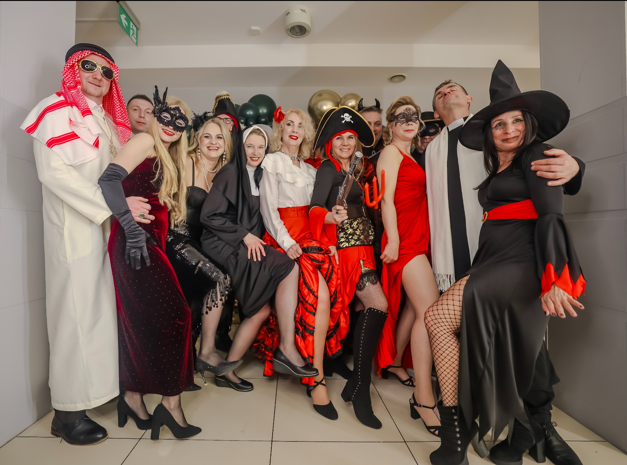 Grupa osób w kostiumach karnawałowych: szejk, zakonnica, piratka, czarownica i inne postacie. Balony w tle. Uśmiechnięte twarze, różnorodne stroje i rekwizyty.
