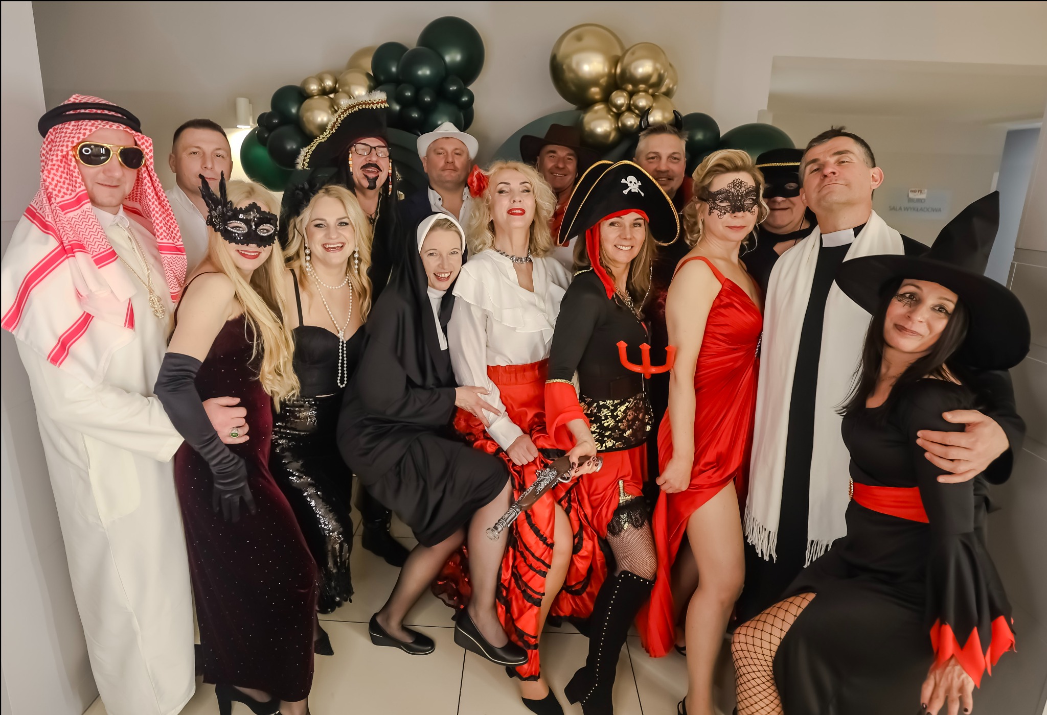 Grupa osób w kostiumach karnawałowych, w tym pirat, szejk, zakonnica i czarownica, pozuje na tle dekoracji z balonów w odcieniach złota i zieleni. Widoczny rekwizyt - zabawkowy pistolet.