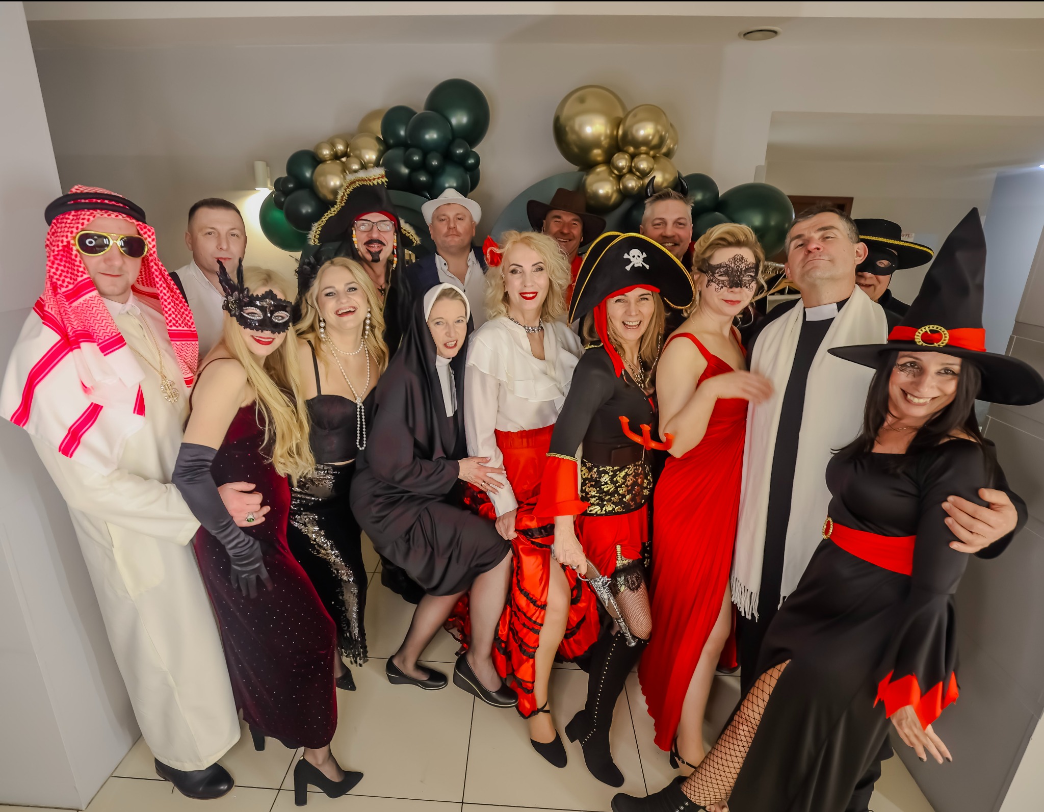 Grupa osób w przebraniach (pirat, szejk, zakonnica, czarownica) pozuje do zdjęcia na tle dekoracji z balonów w odcieniach złota i zieleni. Uśmiechy, kostiumy, imprezowa atmosfera.