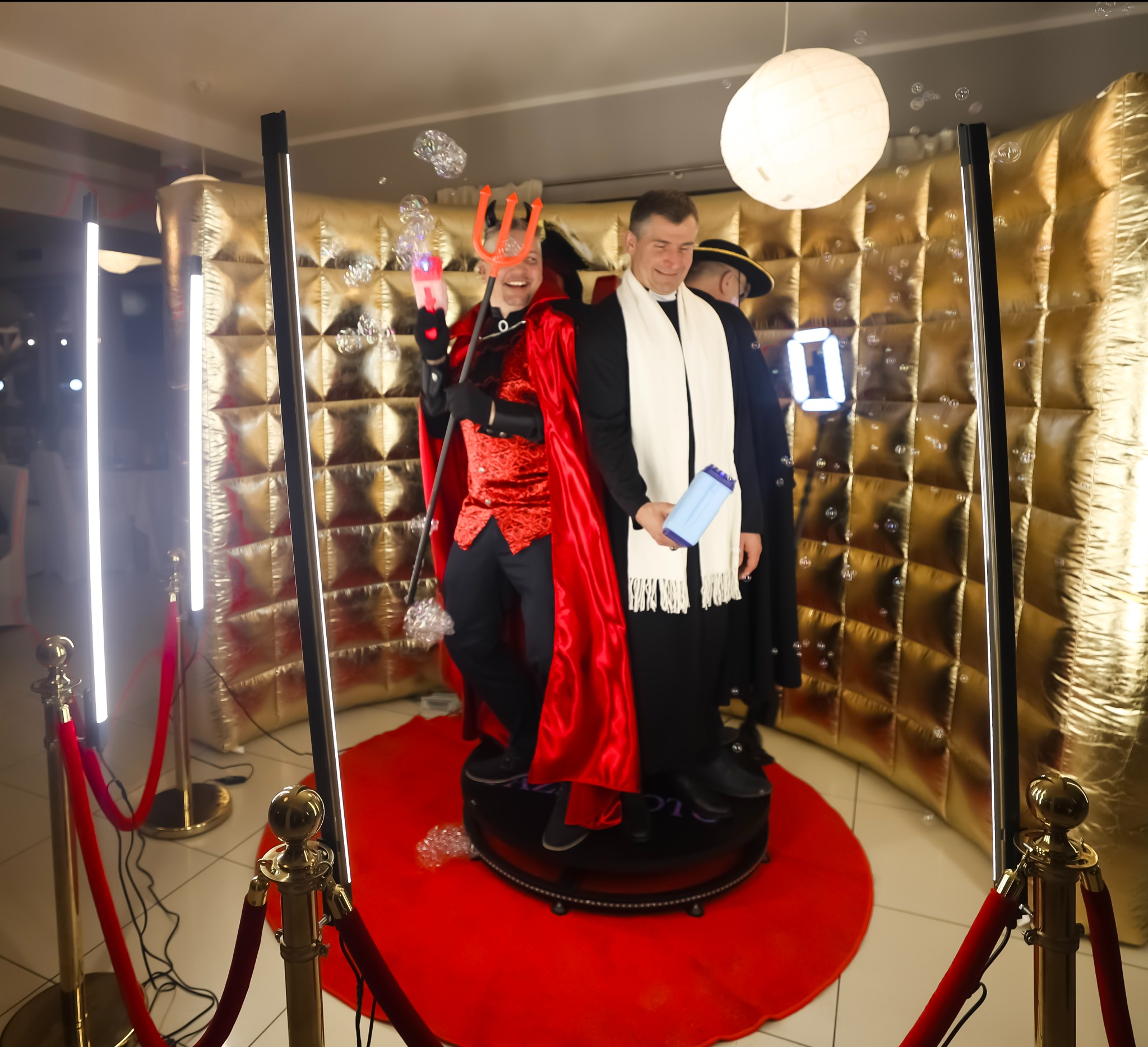 Uśmiechnięci mężczyźni w przebraniach (diabeł i ksiądz) pozują w fotobudce 360 z czerwonym dywanem i złotym tłem. W powietrzu unoszą się bańki mydlane.