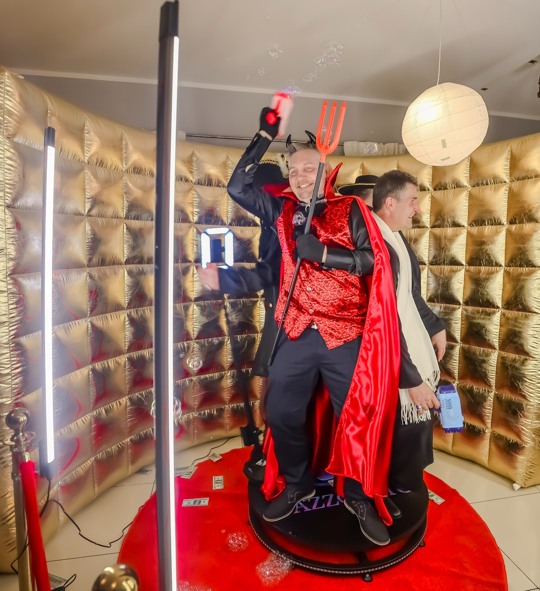 Uczestnicy imprezy w przebraniach pozują w fotobudce z rekwizytami, tworząc radosną atmosferę. Złote tło i czerwony dywan dodają blasku.