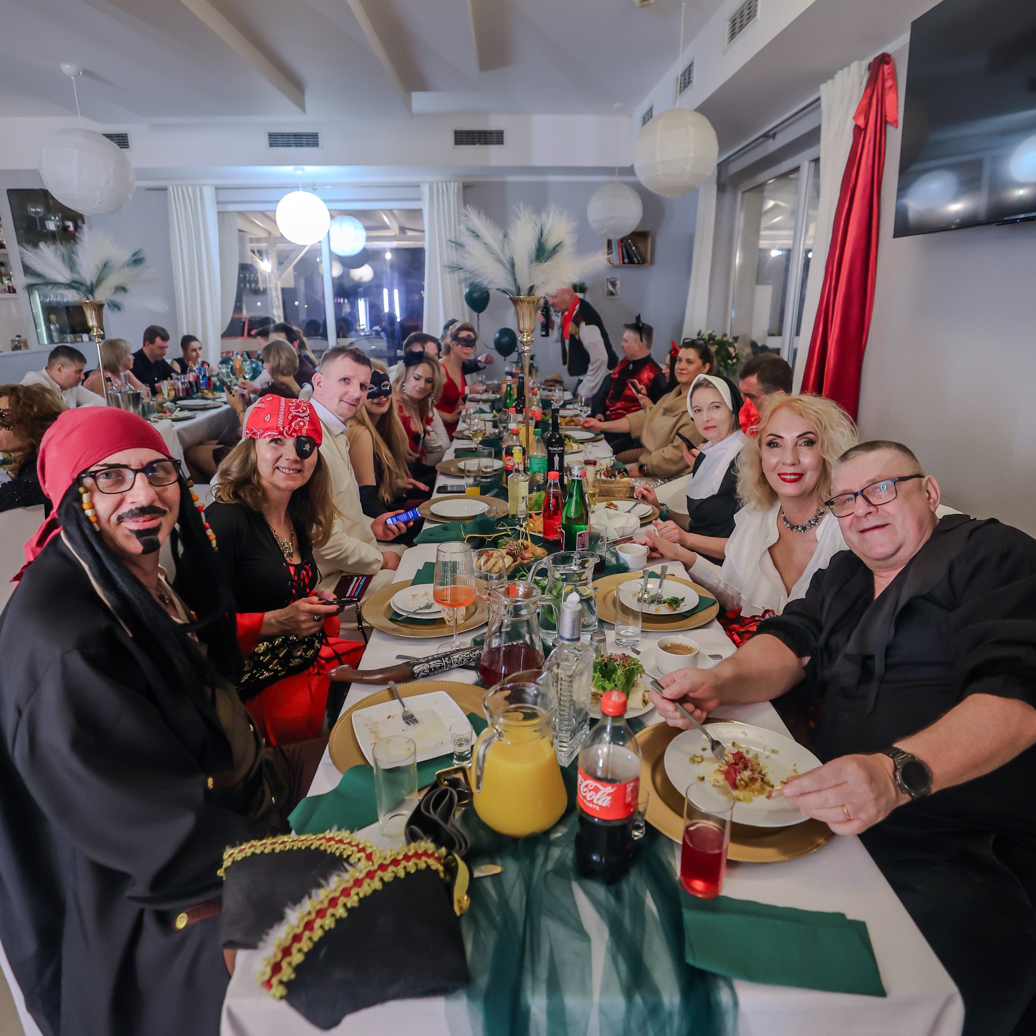 Uczestnicy balu przebierańców w Warszawie siedzą przy stole podczas imprezy. Widoczne dekoracje, napoje i jedzenie. Goście w kostiumach piratów i innych postaci.