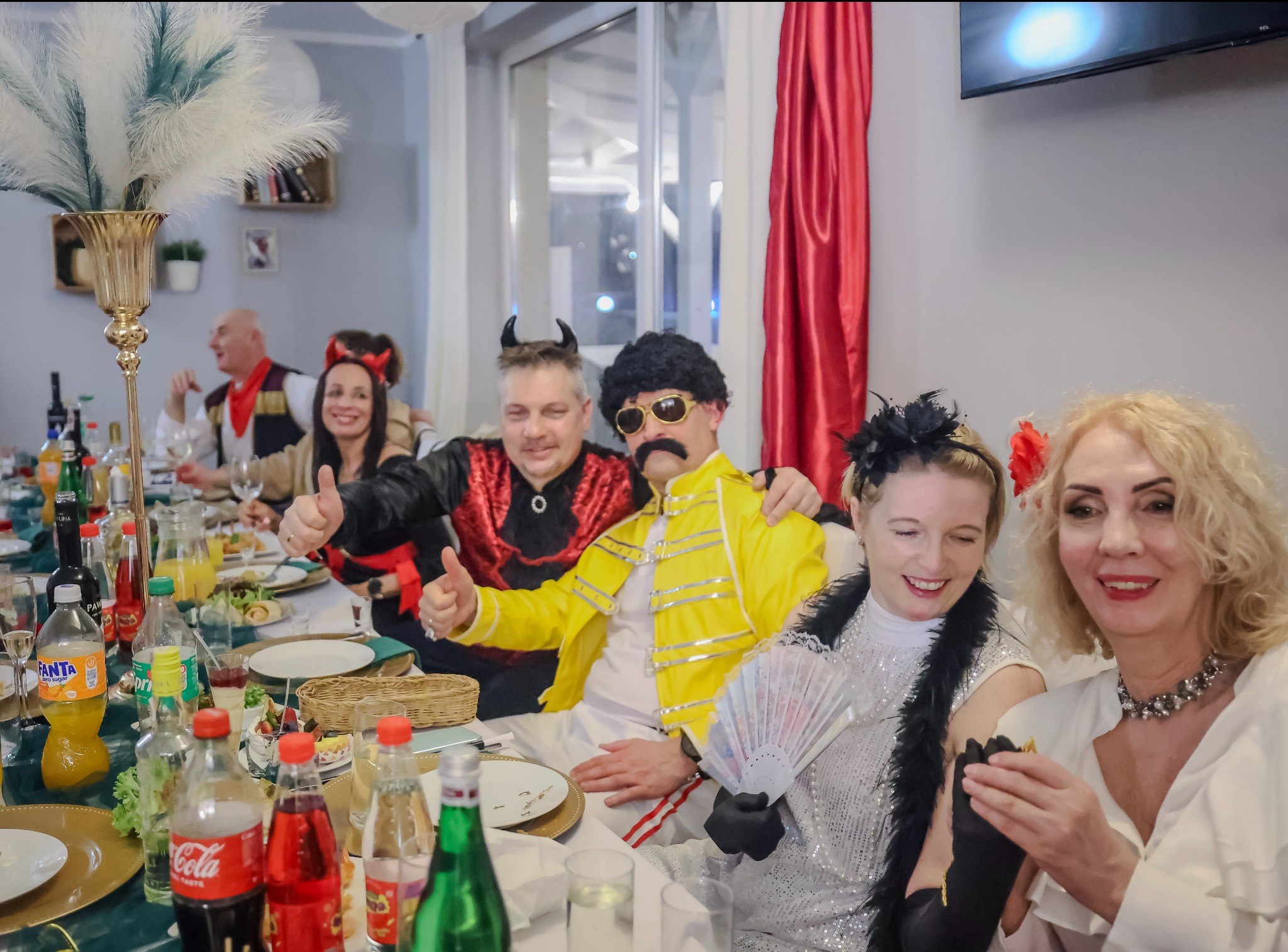 Uczestnicy imprezy tematycznej w kostiumach, siedzący przy stole z napojami i jedzeniem. Widoczny mężczyzna w stroju Freddiego Mercury'ego i kobieta z wachlarzem.