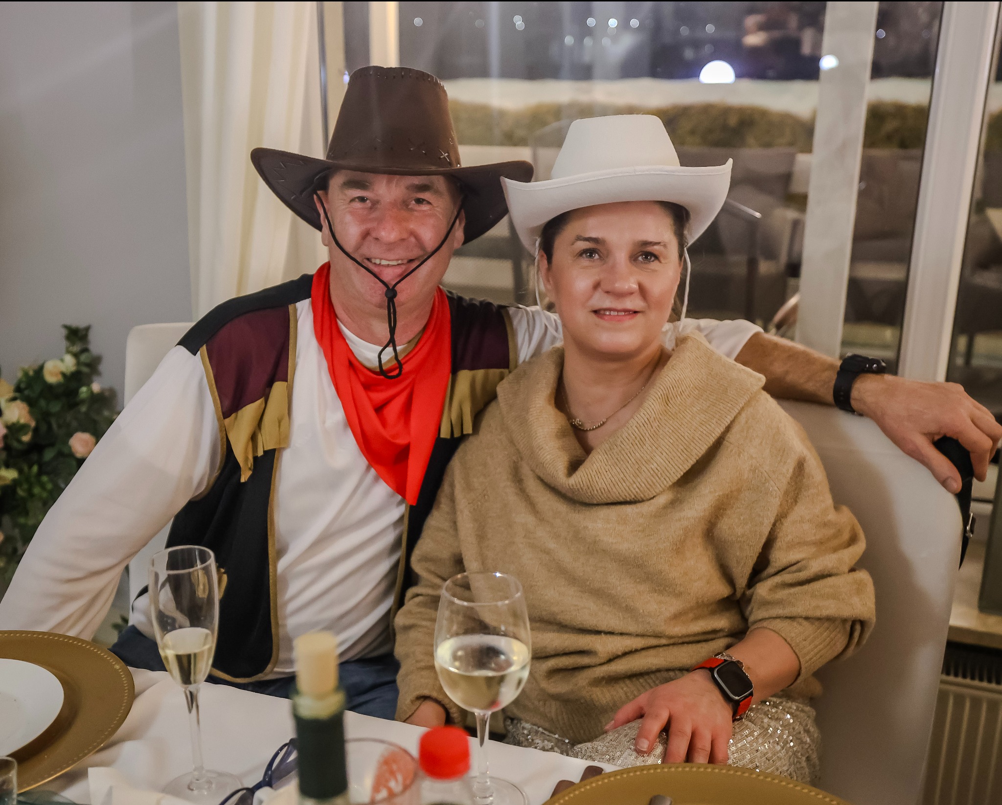 Uśmiechnięta para w strojach kowbojskich siedzi przy stole z kieliszkami wina. Mężczyzna w brązowym kapeluszu i czerwonej chuście, kobieta w białym kapeluszu i beżowym swetrze.