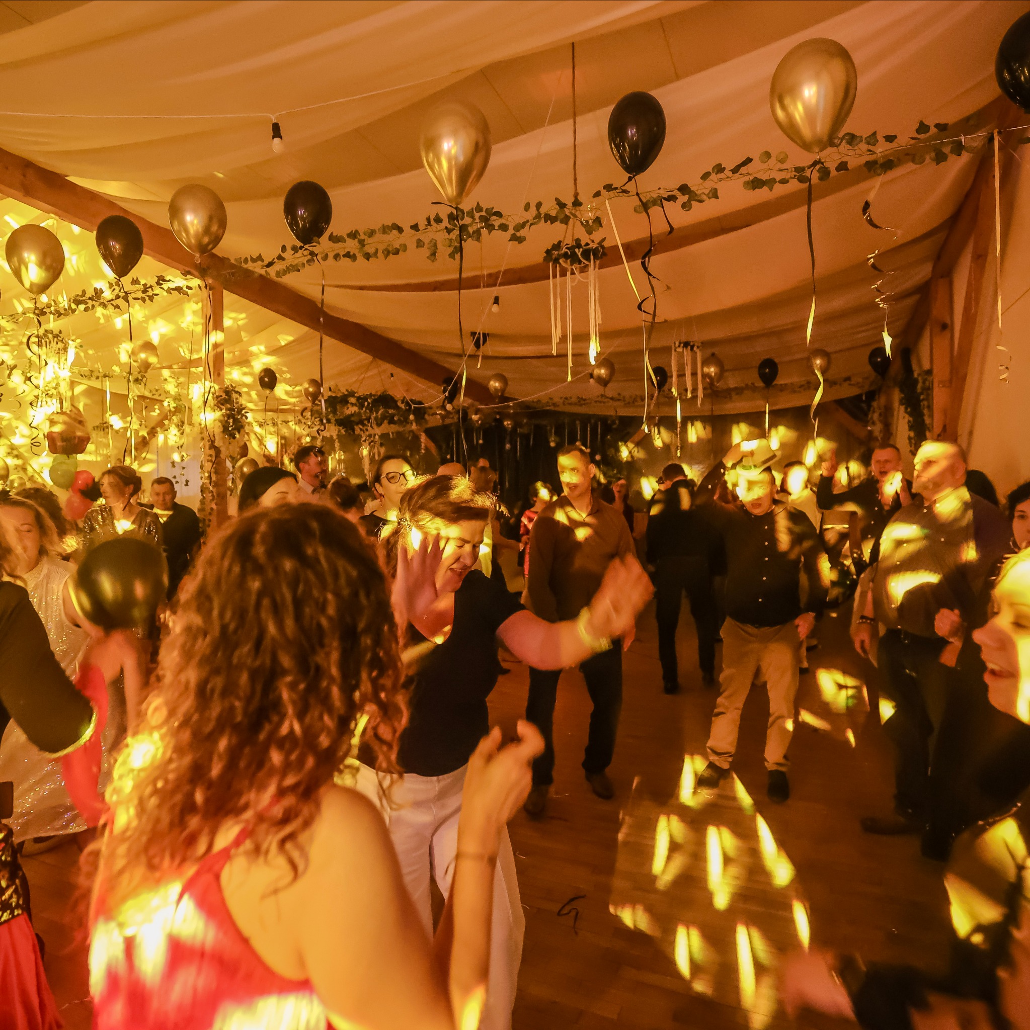 Udekorowana sala balowa z tańczącymi gośćmi, oświetlona ciepłym światłem i ozdobiona balonami w kolorach czerni i złota, tworząca radosną atmosferę imprezy.