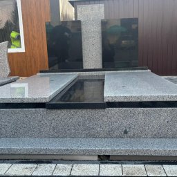 MAT GRAN Nagrobki - Nowoczesny, szary pomnik z czarnymi elementami, ustawiony na kostce brukowej. Widoczne odbicia w polerowanych powierzchniach. W tle budynek z oknem.