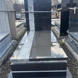 MAT GRAN Nagrobki - Czarny nagrobek z szarym pasem po lewej stronie, na zewnątrz, w tle inne nagrobki i drzewa bez liści. Polerowany kamień odbija niebo.