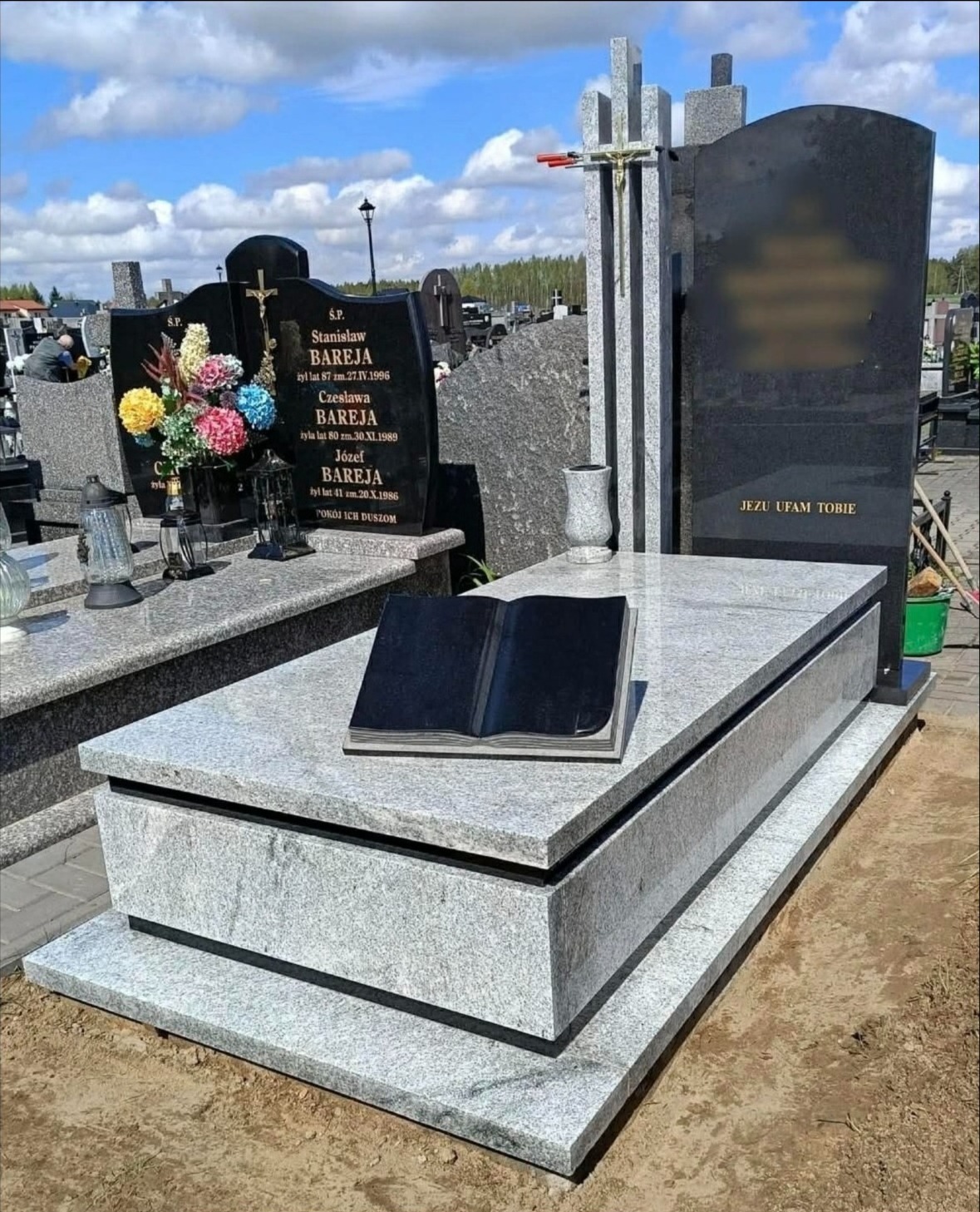 Jasnoszary, granitowy pomnik z czarną, otwartą księgą na płycie. W tle inne pomniki i błękitne niebo z chmurami. Napis 'Jezu ufam Tobie' na pionowej płycie.