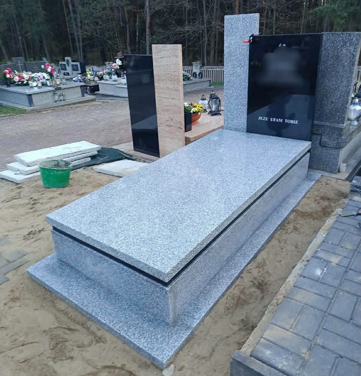 Jasnoszary, granitowy pomnik z czarną tablicą i napisem 'Jezu ufam Tobie', ustawiony na piasku, z widocznymi innymi pomnikami w tle.