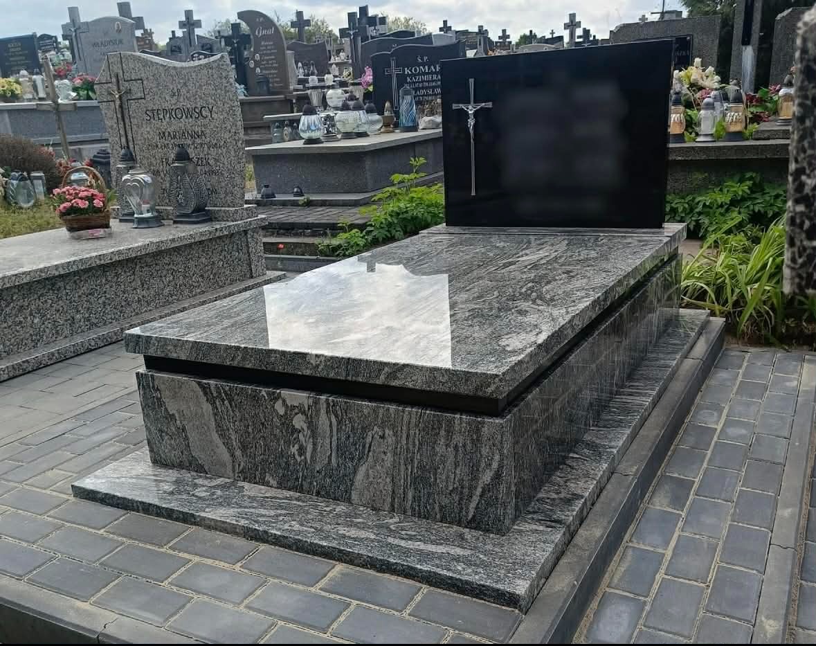 Granitowy nagrobek z czarną tablicą i krzyżem na cmentarzu, otoczony zielenią i innymi nagrobkami. Widok z bliska, podkreślający fakturę kamienia.