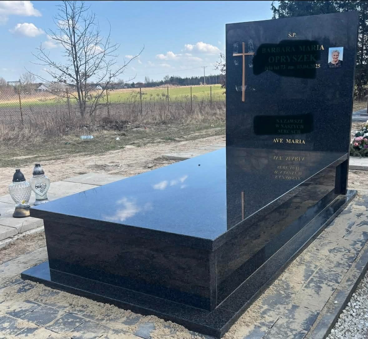 Czarny, polerowany pomnik z granitu z wygrawerowanym napisem i zdjęciem osoby zmarłej, ustawiony na cmentarzu. W tle widoczne pole i drzewa.