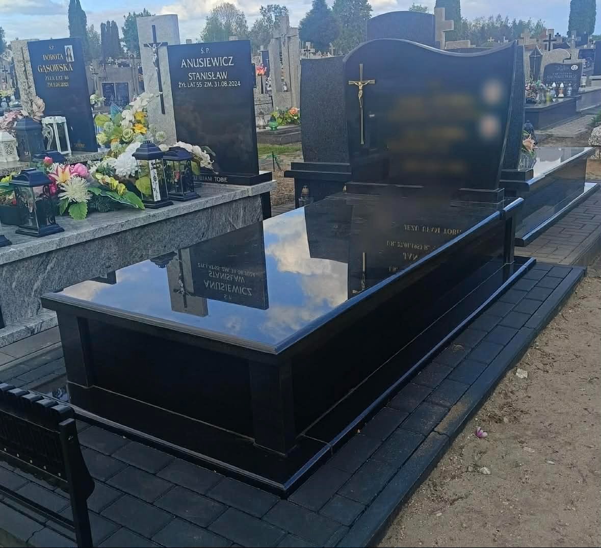 Czarny, polerowany pomnik z krzyżem, odbijający niebo, na cmentarzu. Widoczne inne nagrobki i zieleń. Ujęcie z boku, ukazujące detale wykończenia.