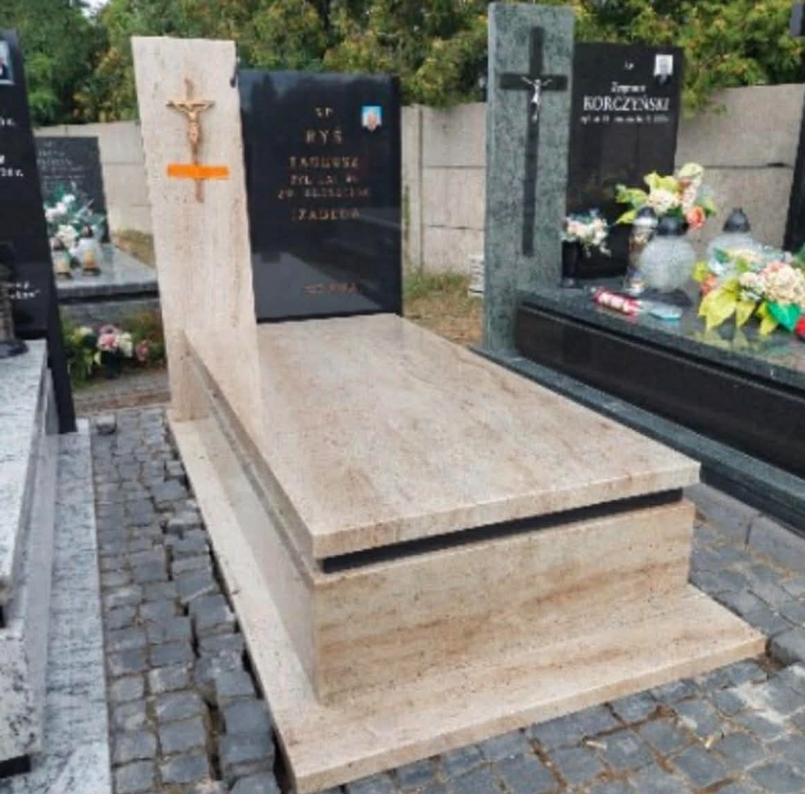 Jasny, beżowy pomnik z krzyżem i czarną tablicą inskrypcyjną, ustawiony na kostce brukowej. Widoczne inne pomniki w tle.