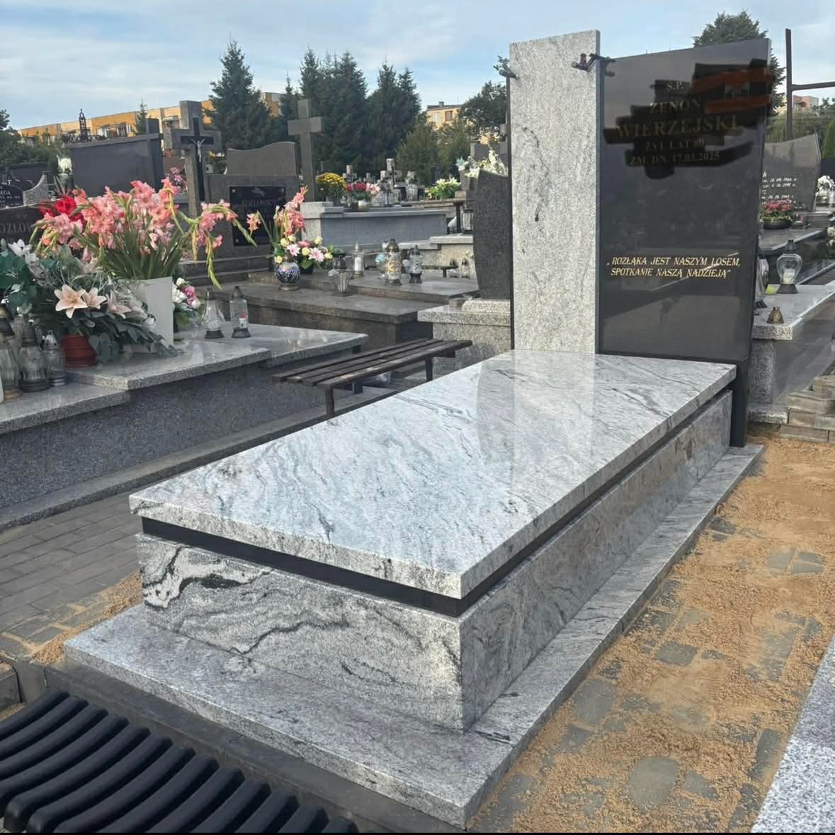 Granitowy pomnik z czarną tablicą inskrypcyjną i jasnoszarym nakryciem, otoczony innymi pomnikami na cmentarzu, w tle drzewa i budynki.