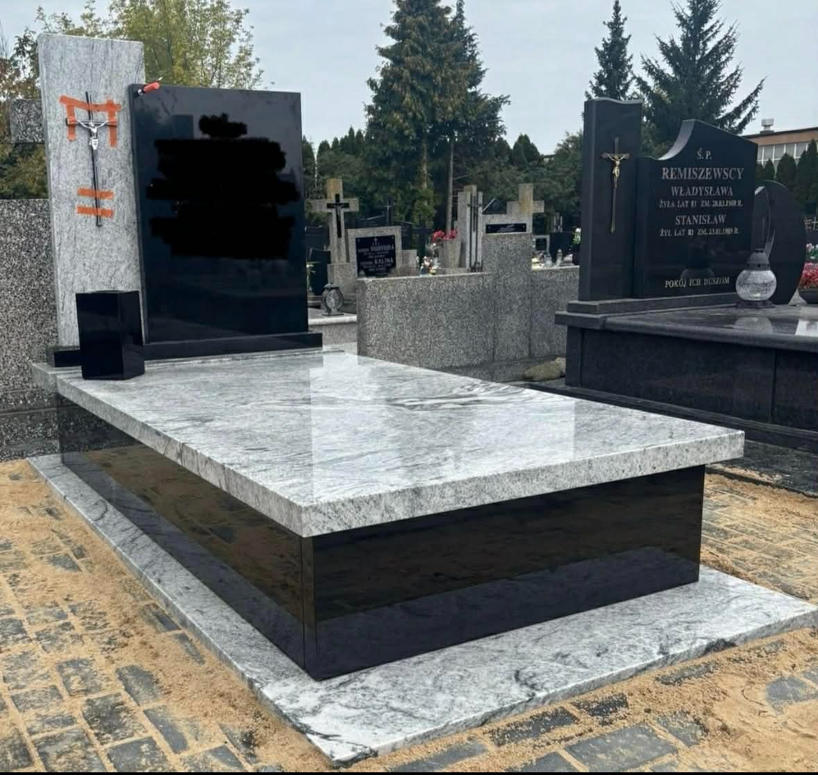 Nowoczesny pomnik z szarego i czarnego granitu na cmentarzu, z krzyżem i tablicą. Widoczne inne groby w tle, drzewa. Całość w stonowanej kolorystyce.