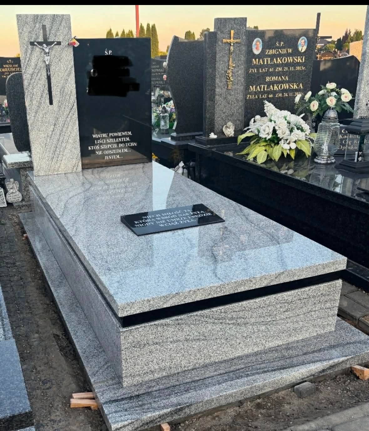 Granitowy grobowiec rodzinny w jasnych odcieniach szarości, z czarnymi tablicami inskrypcyjnymi. Widoczne inne nagrobki w tle, cmentarne otoczenie.