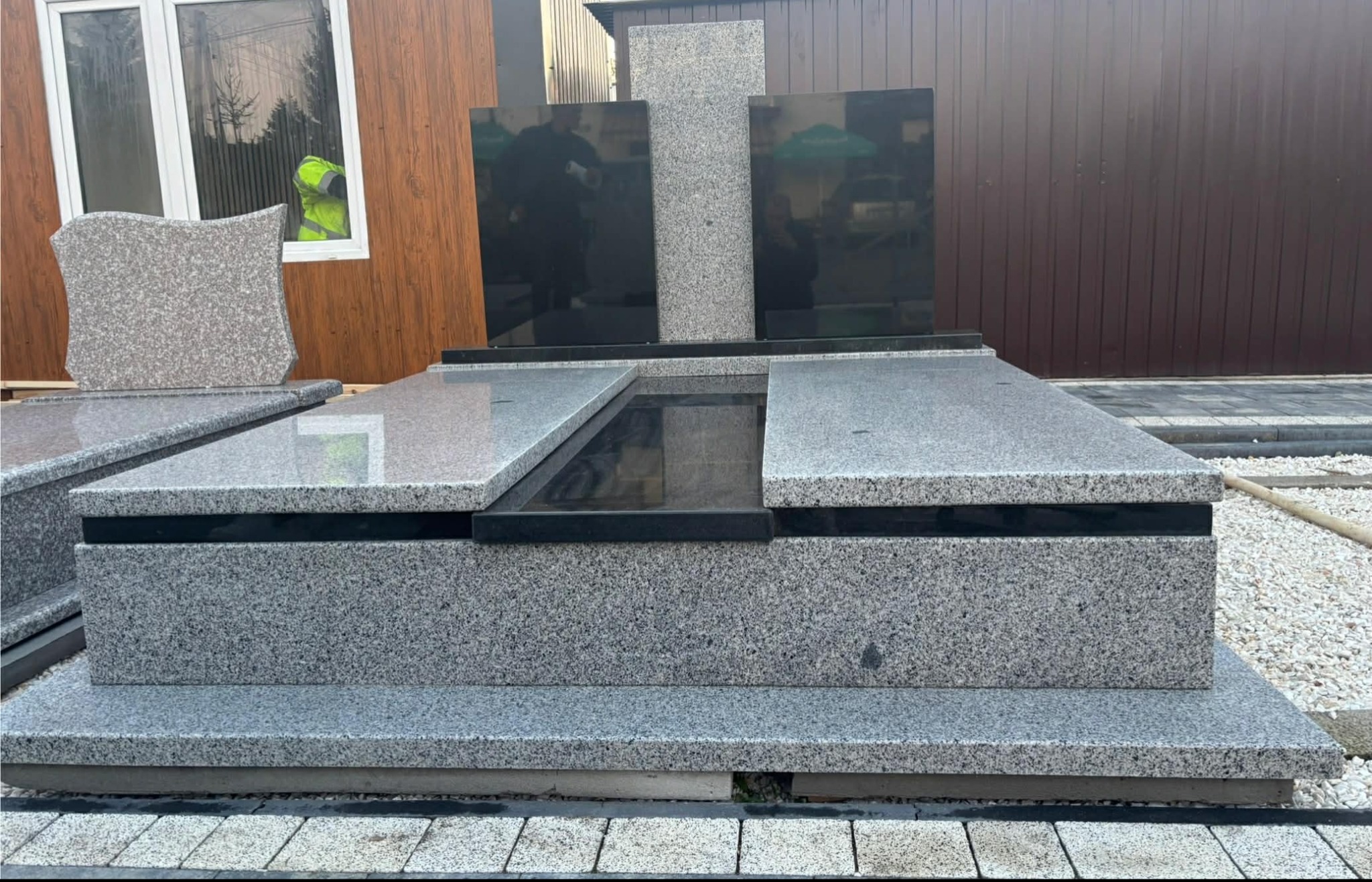 Nowoczesny, szary pomnik z czarnymi elementami, ustawiony na kostce brukowej. Widoczne odbicia w polerowanych powierzchniach. W tle budynek z oknem.