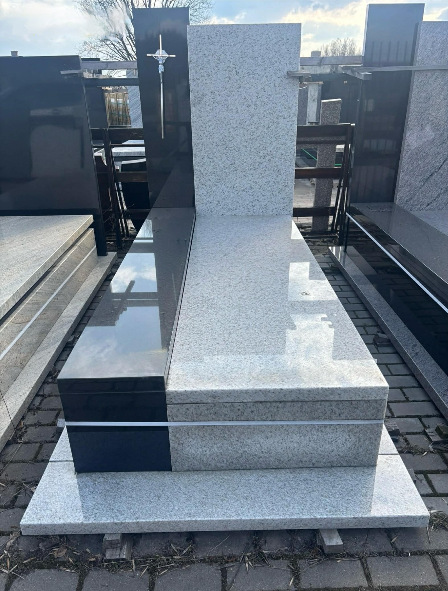 Nowoczesny nagrobek z jasnego granitu z czarnym akcentem i srebrnym krzyżem, ustawiony na kostce brukowej. Widoczne odbicie nieba w polerowanej powierzchni.