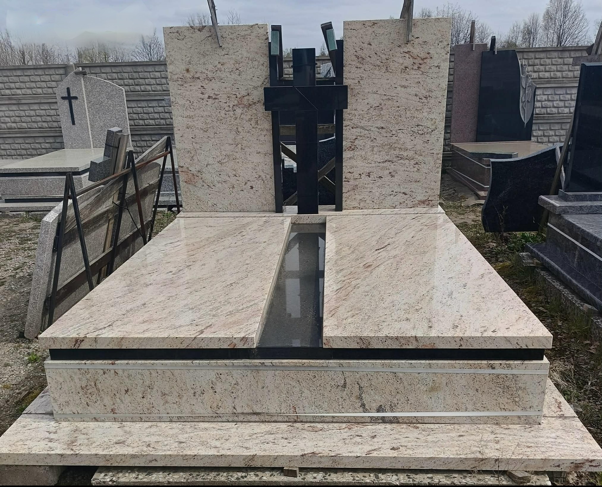 Jasny, granitowy pomnik z czarnym krzyżem w tle, otoczony innymi nagrobkami. Widoczne detale wykończenia i faktura kamienia. Ekspozycja na zewnątrz.