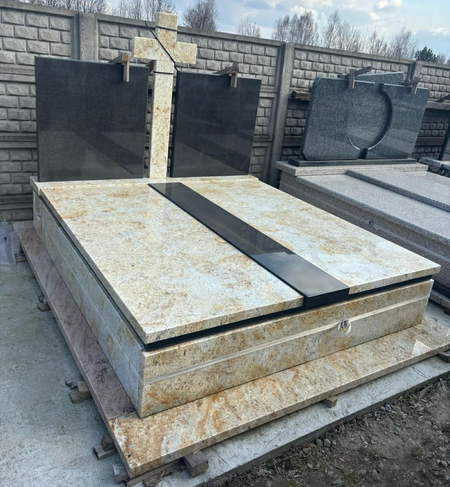 Jasny, granitowy pomnik z ciemnym pasem pośrodku i krzyżem w tle, ustawiony na betonowej podstawie. Widoczne inne nagrobki i betonowe ogrodzenie.