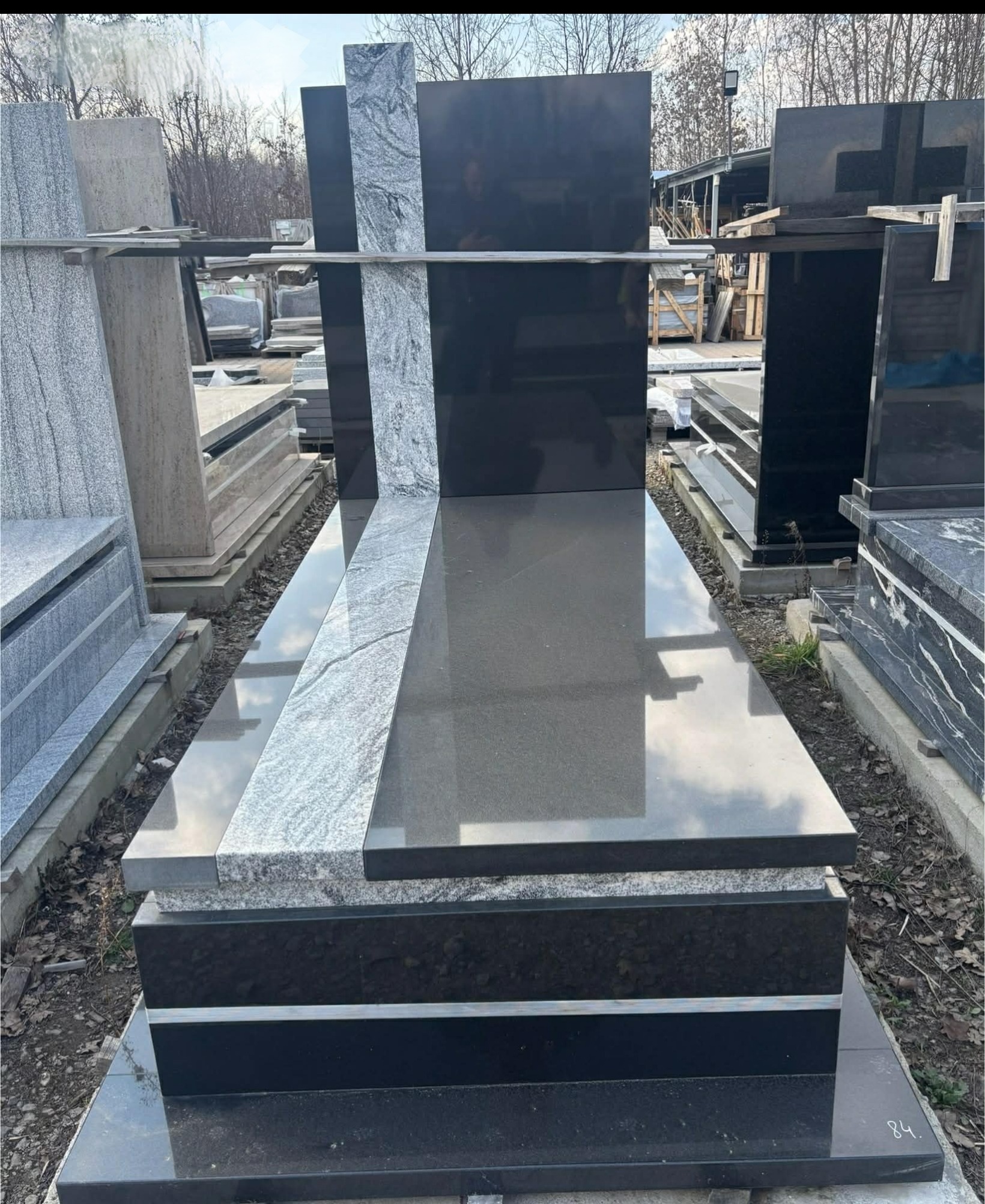 Czarny nagrobek z szarym pasem po lewej stronie, na zewnątrz, w tle inne nagrobki i drzewa bez liści. Polerowany kamień odbija niebo.