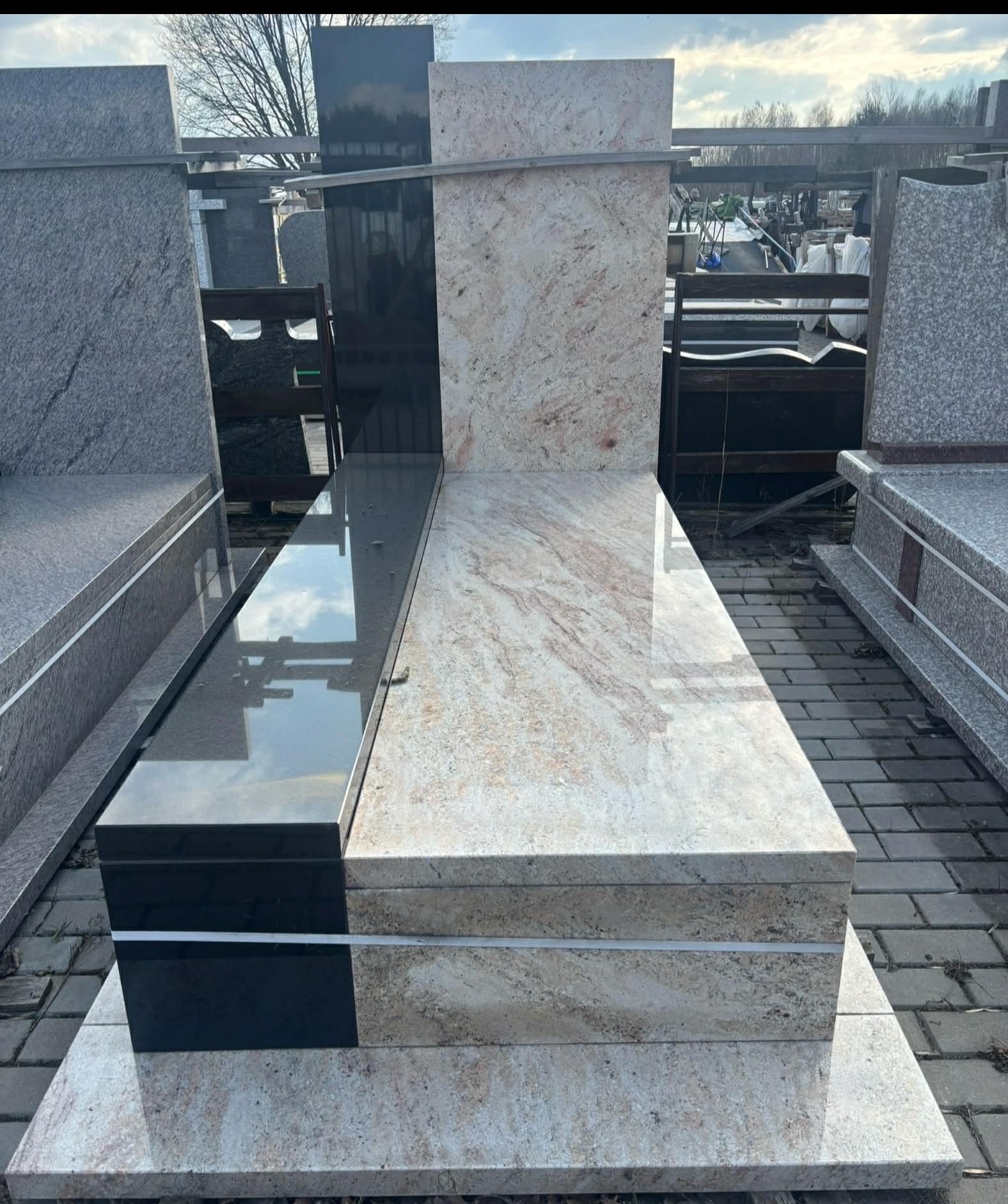 Dwukolorowy, poziomy pomnik z jasnego i czarnego kamienia, z widocznym odbiciem nieba na polerowanej powierzchni. Widoczne inne elementy kamienne w tle.