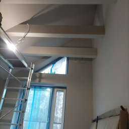 Usługi budowlane Dmytro Krasnikov - Wnętrze w trakcie prac wykończeniowych: ściany po szpachlowaniu, zabezpieczone okna folią, widoczna drabina malarska i drewniane belki pod sufitem.