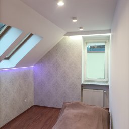Usługi budowlane Dmytro Krasnikov - Pokój na poddaszu z oknem dachowym i wzorzystą tapetą. Podświetlenie LED przy oknie dachowym. Widoczne drewniane panele podłogowe i grzejnik pod oknem.