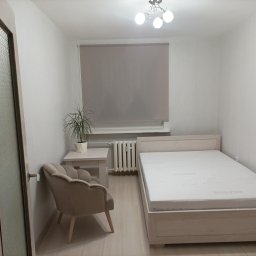 Usługi budowlane Dmytro Krasnikov - Jasne wnętrze sypialni po odświeżeniu: beżowy fotel, łóżko z materacem, roleta w oknie, grzejnik i kwiat doniczkowy na stoliku. Minimalistyczny design, świeżo po remoncie.