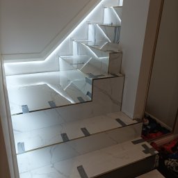 Usługi budowlane Dmytro Krasnikov - Wykończone schody z białego marmuru z podświetleniem LED, zabezpieczone taśmą malarską. Widoczne złote listwy wykończeniowe i odbicie w lustrze z butami.