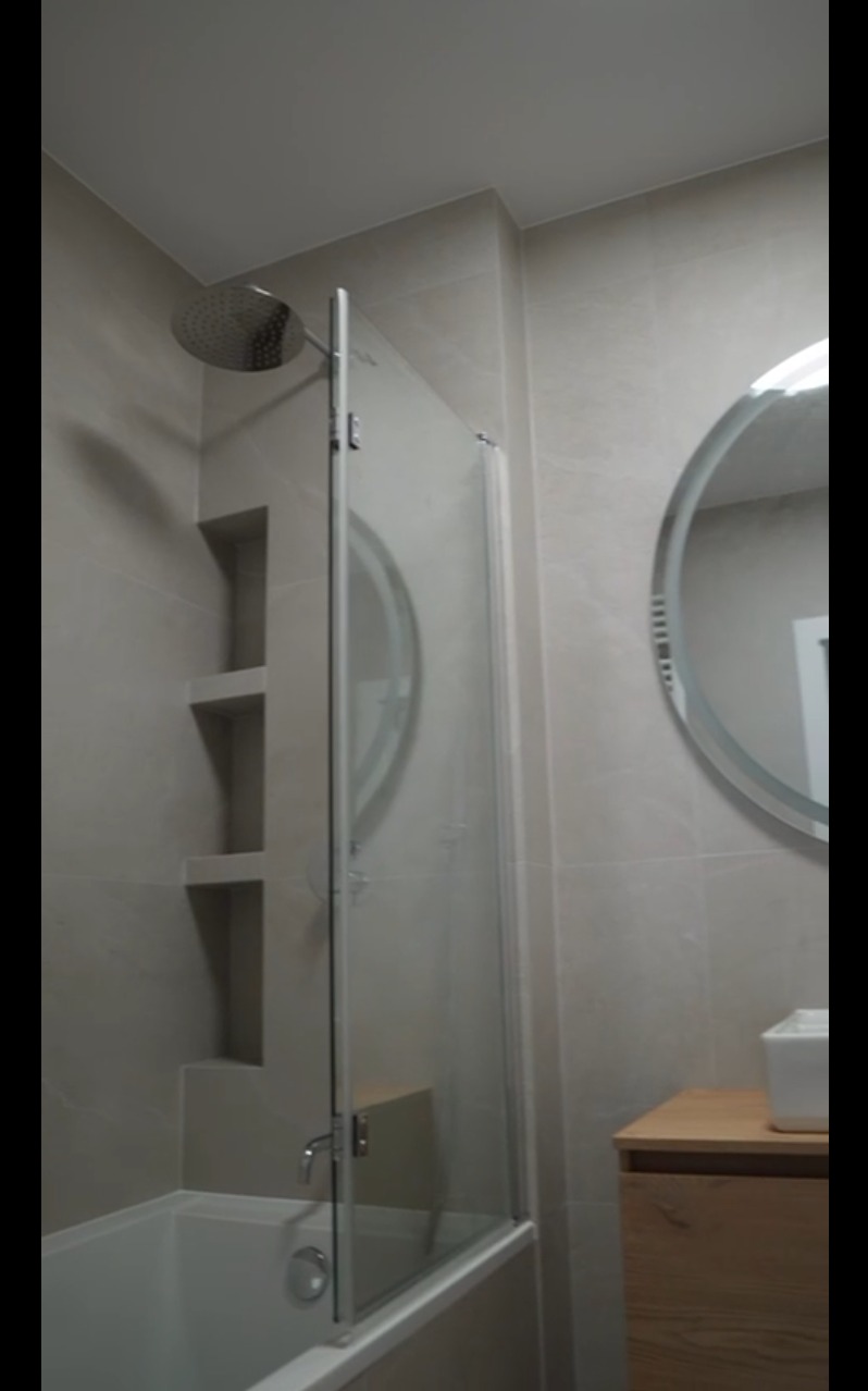 Wykończona łazienka z kabiną prysznicową nad wanną, okrągłym lustrem i wnękami w ścianie wyłożonej jasnymi płytkami. Widoczny fragment szafki pod umywalką.