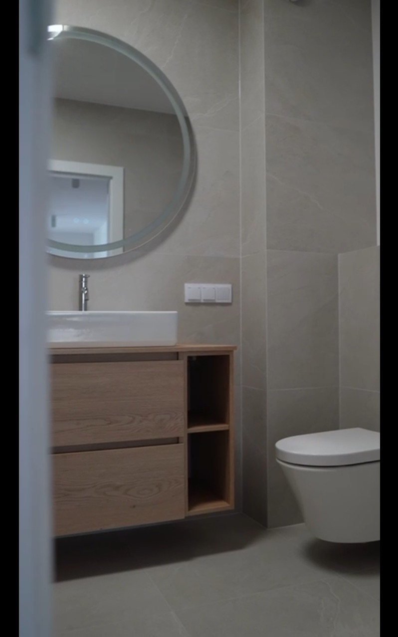 Nowoczesna łazienka z umywalką na szafce z drewna, okrągłym lustrem z podświetleniem LED i podwieszaną toaletą. Szare płytki na ścianach i podłodze.