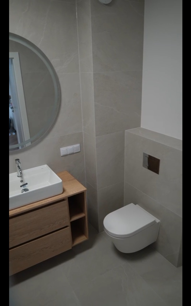 Nowoczesna łazienka z umywalką na drewnianej szafce i podwieszaną toaletą, wykończona szarymi płytkami imitującymi marmur. Okrągłe lustro nad umywalką.